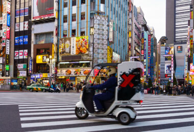 A delivery scooter moves through the large scramble crossing on Central Road in Kabukichō, one of Shinjuku’s busiest commercial corridors. This stretch of the district, located just north of Yasukuni-dōri, has been a dense entertainment zone since the postwar reconstruction era, eventually becoming known for its mix of cinemas, restaurants, arcades, karaoke towers, and neon advertising stacked across narrow mid-rise buildings.

The tall signs and narrow storefronts reflect the vertical commercial pattern typical of Shinjuku’s nightlife blocks, where multiple businesses occupy each floor and compete for visibility at street level. In the distance, the Godzilla head mounted atop the Hotel Gracery Shinjuku—installed in 2015 as part of the Toho Cinemas complex—has become one of the district’s most recognizable landmarks, connecting the area’s contemporary tourism appeal with Japan’s long-running film history.