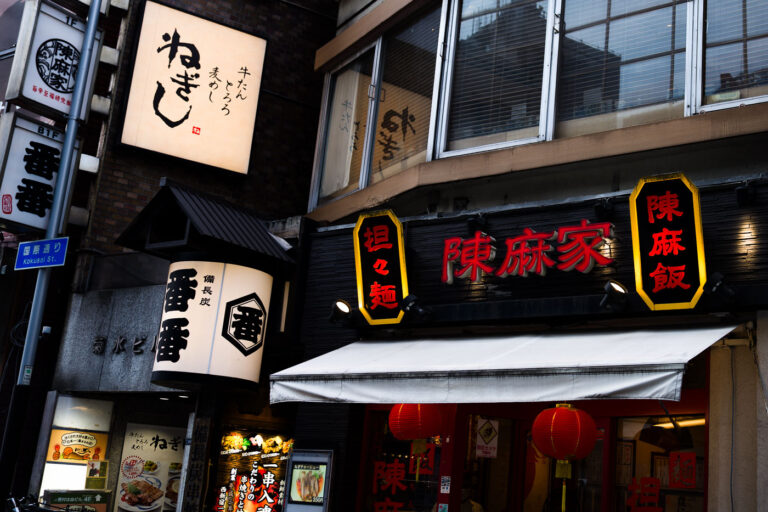 Evening Streetfront in Shinjuku 3 A cluster of restaurants along a narrow Shinjuku street glows with layered signage in both Japanese and Chinese scripts. The red and gold lettering of a Sichuan-style noodle house stands out beneath the muted tones of surrounding izakayas, each sign competing for attention with subtle variations in type and light. Red paper lanterns hang just above the entrance, signaling warmth and spice within, while the larger illuminated kanji above advertise dishes like “tan-tan men” and “mapo rice.” The mix of fonts, textures, and light reflects Tokyo’s dynamic blend of regional cuisines and visual clutter—a dense, living typography of urban appetite and identity.