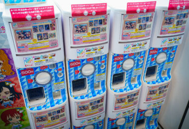 A neat row of colorful Gashapon capsule toy machines stands ready to dispense collectible surprises. Each unit, labeled with the Bandai logo and priced at 500 yen, features themed flat-pack items tied to anime or pop culture series. The bright geometric design and cheerful signage reflect Japan’s mastery of micro-entertainment—where vending and chance intersect to deliver small bursts of joy. These machines, common in train stations and malls, embody a blend of nostalgia, design precision, and everyday whimsy unique to modern Japanese street culture.