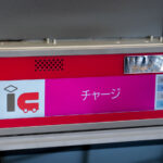 A close-up of a Japanese IC card recharge machine shows a bright pink interface with the word “チャージ” (charge) displayed prominently. The IC logo indicates it’s used for topping up transit smart cards like Suica or Pasmo. On the right side, selectable amounts range from 1,000 to 10,000 yen. The machine’s metal casing and reflective surface hint at its location inside a modern subway or train station.