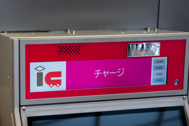 g 3 A close-up of a Japanese IC card recharge machine shows a bright pink interface with the word “チャージ” (charge) displayed prominently. The IC logo indicates it’s used for topping up transit smart cards like Suica or Pasmo. On the right side, selectable amounts range from 1,000 to 10,000 yen. The machine’s metal casing and reflective surface hint at its location inside a modern subway or train station.