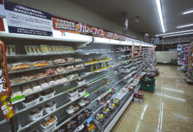 A 7-Eleven convenience store in Japan displays partially empty refrigerated shelves during what appears to be a restocking period or supply shortage. Packaged sandwiches, rice balls, and ready-made curry meals fill the remaining spaces under a banner advertising a “Curry Festival.” The bright fluorescent lighting and orderly layout reflect the precision and consistency typical of Japanese convenience stores, even in moments of limited stock—showing the country’s deep integration of convenience culture into everyday urban life.