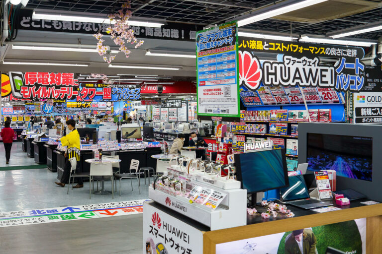 Inside Yodobashi Camera in Shinjuku 3 An interior view of Yodobashi Camera’s sprawling Shinjuku complex in March 2023, showing the densely organized electronics floor where computer hardware, mobile devices, and accessories are displayed under bold, color-saturated signage. The store’s characteristic layout—aisles packed with laptops, desktop components, peripherals, and promotional displays—reflects its role as one of Tokyo’s most comprehensive electronics retailers.Prominent branding for Huawei laptops and smartwatches fills the foreground, while the surrounding aisles feature comparative pricing banners, point-reward offers, and emergency power supply promotions. Yodobashi Camera’s Shinjuku location spans multiple interconnected buildings near the west exit of Shinjuku Station, drawing commuters, hobbyists, and tourists with its high inventory turnover, product variety, and multilingual support.