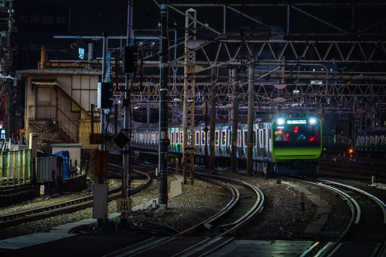 JR East Commuter Train Approaching Station at Night, Japan 3 Illuminated passenger train at night in Japanese railway depot with overhead catenary lines and signal infrastructure.