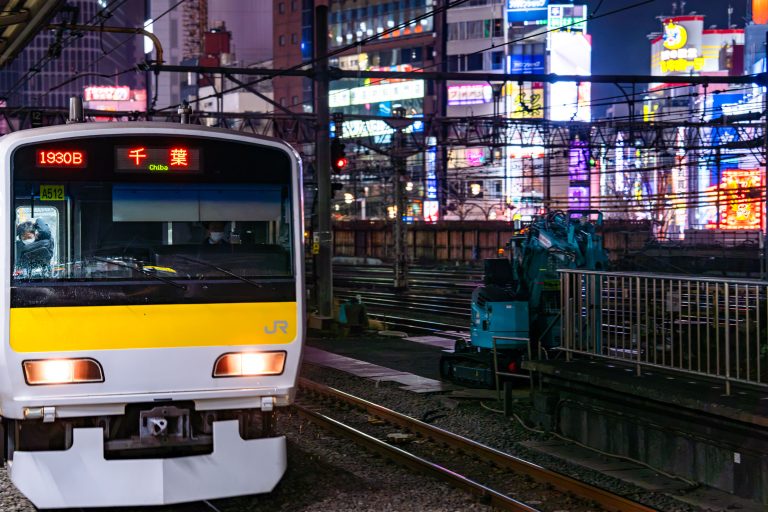 JR East E231 Series Bound for Chiba, Tokyo 1 Yellow and white commuter train stopped at night platform with illuminated city signs behind.