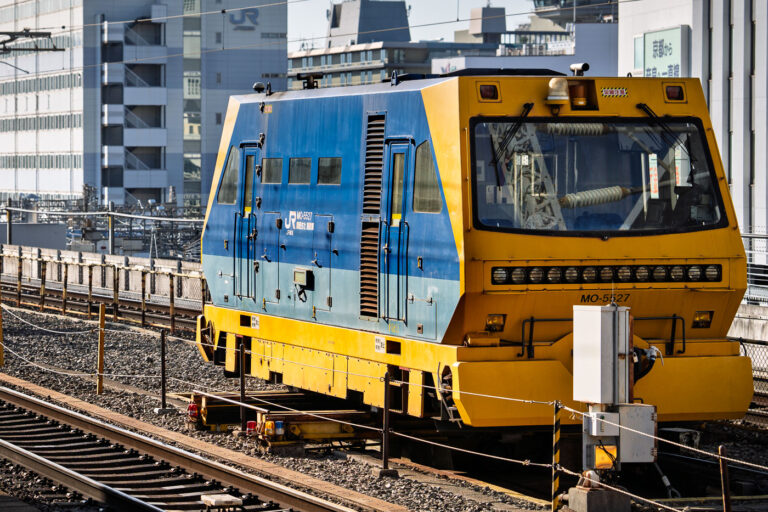 Industrial Maintenance Train, Kyoto 3 A JR West track maintenance vehicle stands on an elevated railway in Kyoto, painted in bright yellow and blue. These machines are vital to Japan’s rail network, used for track inspection, alignment, and upkeep during off-peak hours. The geometric design and industrial strength of the vehicle contrast with the dense urban surroundings, illustrating Japan’s precision engineering and efficiency in rail transport.