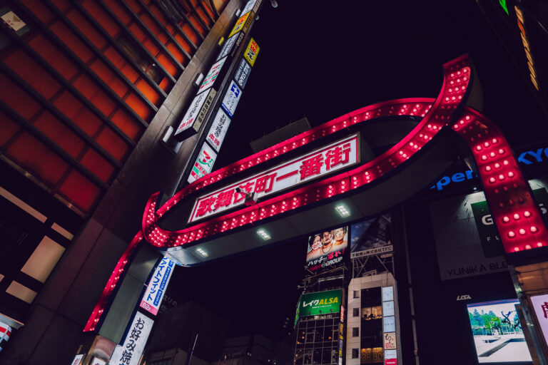 Kabukicho Ichiban-gai Gate at Night 4 The iconic illuminated gate marking Kabukichō Ichiban-gai, photographed in Shinjuku, Tokyo. Installed in the late 1960s and redesigned several times since, the red archway has become one of the most recognizable entrances to Tokyo’s largest entertainment district.
Kabukichō developed after World War II as a planned theater district—its name comes from a never-built kabuki theater—before evolving into a dense zone of restaurants, bars, cinemas, and nightlife venues. The gate’s bright bulbs and bold lettering serve as a symbolic threshold into the neighborhood’s tightly packed streets, which remain active late into the night.