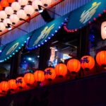 Lantern Light Over Shinjuku Alley 3 A cascade of red and white paper lanterns illuminates the facade of an izakaya in Shinjuku, Tokyo, casting a warm glow against the cool blue tones of the evening. Each lantern is hand-painted with calligraphy, evoking a blend of tradition and nightlife energy that defines Tokyo’s dense entertainment districts. The upper awnings—lined with blue bulbs and framed by wooden beams—hint at the layered complexity of urban Japan, where old-world craftsmanship meets electric color and constant reinvention. The faint reflections of neon and interior lighting merge through the windows, creating a luminous dialogue between the street and the world within.