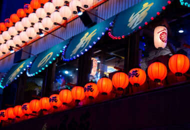 A cascade of red and white paper lanterns illuminates the facade of an izakaya in Shinjuku, Tokyo, casting a warm glow against the cool blue tones of the evening. Each lantern is hand-painted with calligraphy, evoking a blend of tradition and nightlife energy that defines Tokyo’s dense entertainment districts. The upper awnings—lined with blue bulbs and framed by wooden beams—hint at the layered complexity of urban Japan, where old-world craftsmanship meets electric color and constant reinvention. The faint reflections of neon and interior lighting merge through the windows, creating a luminous dialogue between the street and the world within.