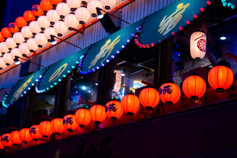Lantern Light Over Shinjuku Alley 2 A cascade of red and white paper lanterns illuminates the facade of an izakaya in Shinjuku, Tokyo, casting a warm glow against the cool blue tones of the evening. Each lantern is hand-painted with calligraphy, evoking a blend of tradition and nightlife energy that defines Tokyo’s dense entertainment districts. The upper awnings—lined with blue bulbs and framed by wooden beams—hint at the layered complexity of urban Japan, where old-world craftsmanship meets electric color and constant reinvention. The faint reflections of neon and interior lighting merge through the windows, creating a luminous dialogue between the street and the world within.