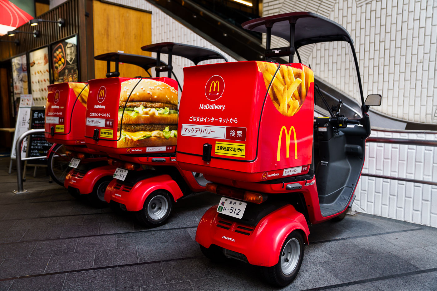 A line of red McDonald's McDelivery scooters, branded with images of a Big Mac and fries, are parked in an urban area in Japan.