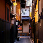 A narrow side alley branching off Hanamikoji-dori in Kyoto’s Gion district, photographed in March 2023. This historic area is known for its preserved machiya townhouses—traditional wooden buildings that date back to the Edo and Meiji periods and once served as both residences and storefronts for merchants and artisans. Many of the structures visible here retain their classic features, including wooden latticework (koshi), earthy plaster walls, deep-eaved roofs, and lanterns marking the entrances of small eateries and teahouses.

Hanamikoji-dori remains closely associated with Kyoto’s hanamachi culture, where geiko and maiko still work in nearby teahouses. While the main street draws steady foot traffic, the smaller lanes behind it—like the one pictured—capture the quieter, more atmospheric side of Gion, where the scale of the architecture narrows and the district’s centuries-old urban fabric becomes especially clear.