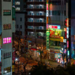 A nighttime view of the commercial streets southwest of Nippori Station in Tokyo’s Arakawa ward, where pachinko parlors, DVD shops, cafés, and convenience stores fill the dense blocks between Nippori and Nishi-Nippori stations. The brightly lit BB Pachinko building and the FamilyMart at street level serve as local landmarks, with narrow side streets branching off toward older residential buildings. The mix of neon signage, small restaurants, and multi-story entertainment businesses reflects the typical streetscape found along the fringe of Tokyo’s major commuter hubs.

This part of Nippori sits just outside the historic Yanaka district, one of the few neighborhoods that survived the 1923 Great Kantō Earthquake and wartime bombing. As the city expanded after the war, the areas around Nippori Station developed into a transit-oriented commercial zone—first serving textile shops and wholesalers, then gradually shifting toward pachinko centers, cafés, and commuter-friendly stores. Today the district remains a blend of old and new Tokyo: dense, walkable, illuminated late into the night, yet still closely connected to the quieter temple districts just a few blocks away.