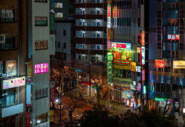 A nighttime view of the commercial streets southwest of Nippori Station in Tokyo’s Arakawa ward, where pachinko parlors, DVD shops, cafés, and convenience stores fill the dense blocks between Nippori and Nishi-Nippori stations. The brightly lit BB Pachinko building and the FamilyMart at street level serve as local landmarks, with narrow side streets branching off toward older residential buildings. The mix of neon signage, small restaurants, and multi-story entertainment businesses reflects the typical streetscape found along the fringe of Tokyo’s major commuter hubs.

This part of Nippori sits just outside the historic Yanaka district, one of the few neighborhoods that survived the 1923 Great Kantō Earthquake and wartime bombing. As the city expanded after the war, the areas around Nippori Station developed into a transit-oriented commercial zone—first serving textile shops and wholesalers, then gradually shifting toward pachinko centers, cafés, and commuter-friendly stores. Today the district remains a blend of old and new Tokyo: dense, walkable, illuminated late into the night, yet still closely connected to the quieter temple districts just a few blocks away.