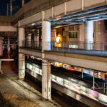 A commuter train passes through Nippori Station in Tokyo beneath an elevated expressway. Nippori is a major interchange for the JR Yamanote, Keisei, and Joban lines, connecting central Tokyo with Narita Airport and surrounding neighborhoods. The scene reflects the district’s tightly integrated infrastructure, where multiple rail lines and highways converge within a compact urban corridor.