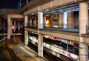 A commuter train passes through Nippori Station in Tokyo beneath an elevated expressway. Nippori is a major interchange for the JR Yamanote, Keisei, and Joban lines, connecting central Tokyo with Narita Airport and surrounding neighborhoods. The scene reflects the district’s tightly integrated infrastructure, where multiple rail lines and highways converge within a compact urban corridor.