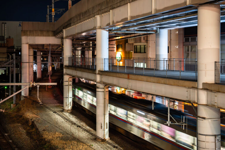 Nippori Station at night 2 A commuter train passes through Nippori Station in Tokyo beneath an elevated expressway. Nippori is a major interchange for the JR Yamanote, Keisei, and Joban lines, connecting central Tokyo with Narita Airport and surrounding neighborhoods. The scene reflects the district’s tightly integrated infrastructure, where multiple rail lines and highways converge within a compact urban corridor.
