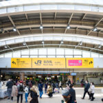 Passengers move through the North Gate of JR Nippori Station, a key interchange for the Yamanote, Keihin-Tohoku, and Joban lines as well as the Nippori–Toneri Liner. Prominent yellow JR East signage directs travelers toward the West Exit for Yanaka and the East Exit for Ueno. The open, modern concourse design reflects Nippori’s role as both a commuter hub and gateway to nearby traditional neighborhoods.