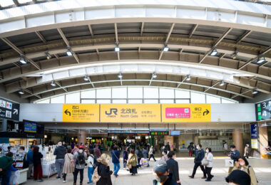 Passengers move through the North Gate of JR Nippori Station, a key interchange for the Yamanote, Keihin-Tohoku, and Joban lines as well as the Nippori–Toneri Liner. Prominent yellow JR East signage directs travelers toward the West Exit for Yanaka and the East Exit for Ueno. The open, modern concourse design reflects Nippori’s role as both a commuter hub and gateway to nearby traditional neighborhoods.