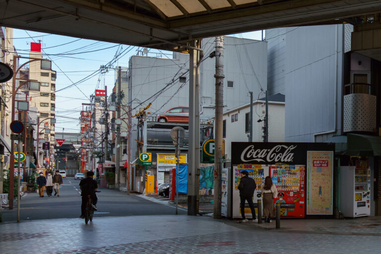 Osaka Street with Coca-Cola Vending Machine 1 A street in Osaka, Japan, with a Coca-Cola vending machine displaying a local map. The urban environment features typical Japanese architecture and overhead wires.
