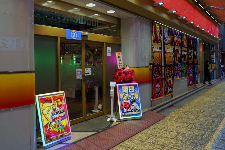 Pachinko Parlor Entrance in Shinjuku 2 Pachinko parlors remain a defining part of Japan’s postwar entertainment landscape, evolving from small mechanical game halls into large, brightly lit venues that line the streets of major cities. This Shinjuku location reflects the modern industry’s mix of gambling-adjacent gaming, animated advertising, and elaborate storefront displays designed to attract commuters and tourists.Although pachinko operates in a legal gray zone—winnings are exchanged off-site to comply with gambling regulations—it has grown into a multibillion-yen sector with deep cultural roots, from the machines’ lineage in early 20th-century children’s games to their present role as a staple of urban nightlife.