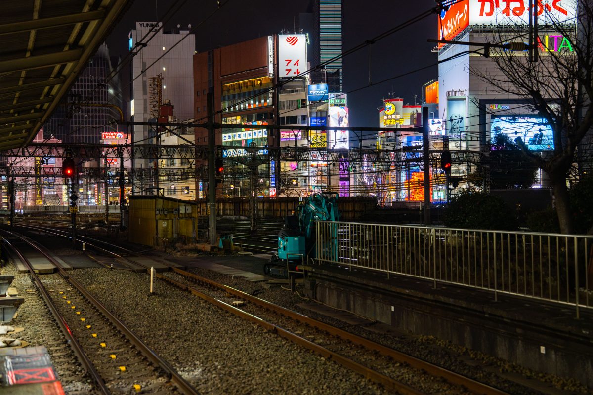 Train tracks and platform at night with illuminated neon signs and buildings in urban Japanese setting.