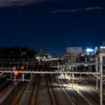 Multiple rail lines converge near Nippori Station in Tokyo, a key junction connecting the JR Yamanote, Keisei, and Joban lines. The illuminated signals and overhead power lines reveal the complexity of the city’s rail infrastructure as it weaves through residential and industrial zones. To the right, elevated expressway supports and nearby buildings frame the corridor, emphasizing Tokyo’s dense layering of transport systems and urban development.