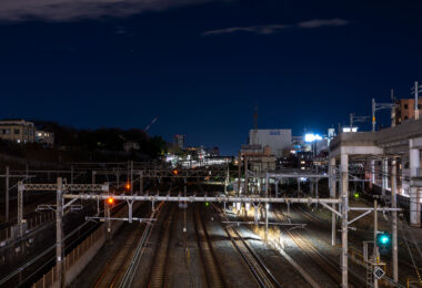 Multiple rail lines converge near Nippori Station in Tokyo, a key junction connecting the JR Yamanote, Keisei, and Joban lines. The illuminated signals and overhead power lines reveal the complexity of the city’s rail infrastructure as it weaves through residential and industrial zones. To the right, elevated expressway supports and nearby buildings frame the corridor, emphasizing Tokyo’s dense layering of transport systems and urban development.