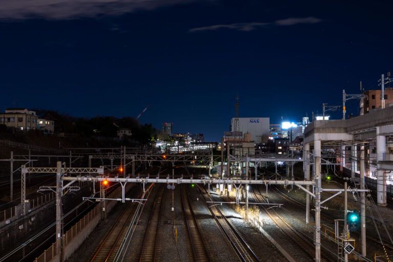 Rails outside Nippori Station in Tokyo 3 Multiple rail lines converge near Nippori Station in Tokyo, a key junction connecting the JR Yamanote, Keisei, and Joban lines. The illuminated signals and overhead power lines reveal the complexity of the city’s rail infrastructure as it weaves through residential and industrial zones. To the right, elevated expressway supports and nearby buildings frame the corridor, emphasizing Tokyo’s dense layering of transport systems and urban development.