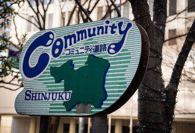 An older enamel “Community Road” sign in Shinjuku, Tokyo, photographed in March 2023. These signs—marked with コミュニティ道路 (“community road”) and a stylized map of the ward—were installed beginning in the late 1970s and 1980s as part of a municipal effort to identify quieter local streets prioritized for pedestrians, schools, and neighborhood traffic rather than through-traffic.

Although many have faded or disappeared as Shinjuku redeveloped, surviving signs like this one remain a small reminder of earlier phases of the ward’s streetscape planning. The hand-drawn lettering, green stripes, and simplified ward outline reflect the graphic design style common in Tokyo’s municipal signage of that era.
