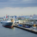 A cargo ship is docked at an industrial pier surrounded by warehouses and factory buildings, their signs bearing familiar Japanese corporate logos. The port’s calm waters and moored vessels hint at the steady rhythm of maritime logistics, with goods flowing in and out of Japan’s manufacturing heartlands. Overhead, a muted sky and the ship’s red funnel add contrast to the subdued industrial palette, capturing a typical day in one of the nation’s bustling coastal trade zones.