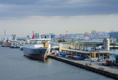 A cargo ship is docked at an industrial pier surrounded by warehouses and factory buildings, their signs bearing familiar Japanese corporate logos. The port’s calm waters and moored vessels hint at the steady rhythm of maritime logistics, with goods flowing in and out of Japan’s manufacturing heartlands. Overhead, a muted sky and the ship’s red funnel add contrast to the subdued industrial palette, capturing a typical day in one of the nation’s bustling coastal trade zones.