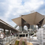 Tokyo Big Sight—officially the Tokyo International Exhibition Center—stands out with its striking inverted-pyramid conference towers, a landmark of the Odaiba and Ariake waterfront districts since its opening in 1996. Designed by architect AXS Satow, the building’s geometric titanium-clad forms and elevated walkways embody the futuristic aesthetic that defined Tokyo’s 1990s bay-area redevelopment. The elevated pedestrian route shown here leads toward the main entrance, filled with visitors streaming in for one of the venue’s frequent trade fairs, expos, and cultural events.

The convention center was built as part of a sweeping effort to revitalize reclaimed land along Tokyo Bay, transforming former industrial zones into hubs for technology, design, and international commerce. Its proximity to the automated Yurikamome transit line helped anchor Odaiba as a showcase for modern urban planning, connecting attractions like Rainbow Bridge, Fuji TV, and the waterfront parks. Today, Tokyo Big Sight remains one of Japan’s busiest exhibition complexes and a defining piece of the city’s contemporary architectural heritage.