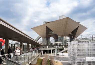 Tokyo Big Sight—officially the Tokyo International Exhibition Center—stands out with its striking inverted-pyramid conference towers, a landmark of the Odaiba and Ariake waterfront districts since its opening in 1996. Designed by architect AXS Satow, the building’s geometric titanium-clad forms and elevated walkways embody the futuristic aesthetic that defined Tokyo’s 1990s bay-area redevelopment. The elevated pedestrian route shown here leads toward the main entrance, filled with visitors streaming in for one of the venue’s frequent trade fairs, expos, and cultural events.

The convention center was built as part of a sweeping effort to revitalize reclaimed land along Tokyo Bay, transforming former industrial zones into hubs for technology, design, and international commerce. Its proximity to the automated Yurikamome transit line helped anchor Odaiba as a showcase for modern urban planning, connecting attractions like Rainbow Bridge, Fuji TV, and the waterfront parks. Today, Tokyo Big Sight remains one of Japan’s busiest exhibition complexes and a defining piece of the city’s contemporary architectural heritage.