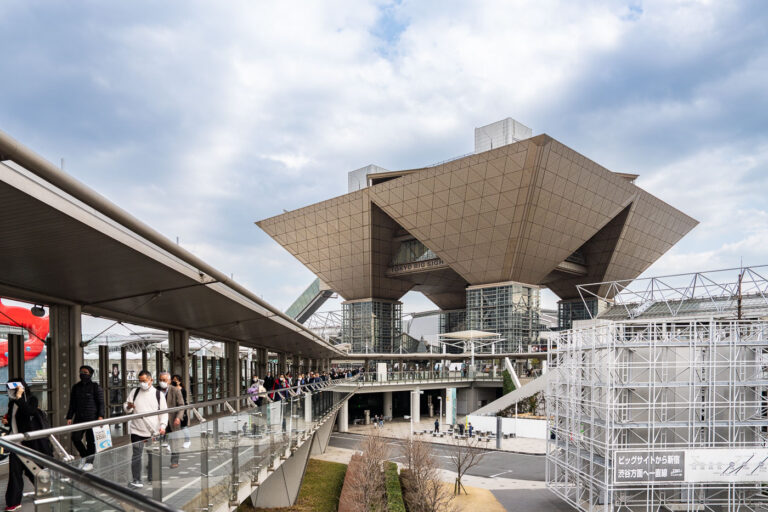 Tokyo Big Sight, Ariake — February 2024 2 Tokyo Big Sight—officially the Tokyo International Exhibition Center—stands out with its striking inverted-pyramid conference towers, a landmark of the Odaiba and Ariake waterfront districts since its opening in 1996. Designed by architect AXS Satow, the building’s geometric titanium-clad forms and elevated walkways embody the futuristic aesthetic that defined Tokyo’s 1990s bay-area redevelopment. The elevated pedestrian route shown here leads toward the main entrance, filled with visitors streaming in for one of the venue’s frequent trade fairs, expos, and cultural events.The convention center was built as part of a sweeping effort to revitalize reclaimed land along Tokyo Bay, transforming former industrial zones into hubs for technology, design, and international commerce. Its proximity to the automated Yurikamome transit line helped anchor Odaiba as a showcase for modern urban planning, connecting attractions like Rainbow Bridge, Fuji TV, and the waterfront parks. Today, Tokyo Big Sight remains one of Japan’s busiest exhibition complexes and a defining piece of the city’s contemporary architectural heritage.