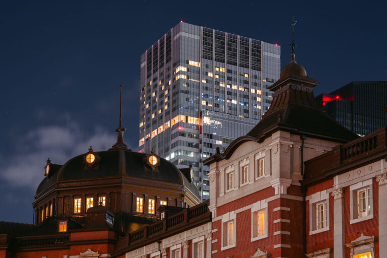 Tokyo Station Marunouchi Facade at Dusk 2 The historic Marunouchi side of Tokyo Station glows warmly at dusk, its red-brick exterior and copper domes standing in contrast to the modern skyscrapers of the surrounding Marunouchi business district. Opened in 1914 and designed by architect Tatsuno Kingo, the station served as the central gateway for Japan’s expanding rail network during the early 20th century.
The dramatic juxtaposition in this March 2023 photograph highlights more than a century of architectural evolution: the restored station building—meticulously reconstructed after wartime damage—foregrounds the sleek high-rise towers that define contemporary central Tokyo. Together, they illustrate how Tokyo layers historic preservation and modern development within one of the busiest transport hubs in the world.