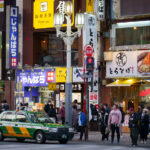 A Tokyo taxi waits at a crossing in Kabukichō, the entertainment district of Shinjuku known for its dense concentration of restaurants, bars, and late-night businesses. The area around Yasukuni-dōri and Shinjuku Station’s east side has long been one of Tokyo’s busiest pedestrian zones, with a mix of small eateries, electronics resellers, and long-established cafés occupying the mid-rise commercial blocks.

The green-and-yellow taxi livery seen here is one of Tokyo’s standard color schemes, used by several of the city’s major cab companies since the late twentieth century. The surrounding storefronts reflect Kabukichō’s layered commercial history, where postwar restaurants, national chains, and contemporary pop-culture signage coexist within the same streetscape.