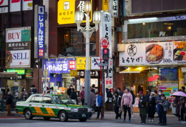 A Tokyo taxi waits at a crossing in Kabukichō, the entertainment district of Shinjuku known for its dense concentration of restaurants, bars, and late-night businesses. The area around Yasukuni-dōri and Shinjuku Station’s east side has long been one of Tokyo’s busiest pedestrian zones, with a mix of small eateries, electronics resellers, and long-established cafés occupying the mid-rise commercial blocks.

The green-and-yellow taxi livery seen here is one of Tokyo’s standard color schemes, used by several of the city’s major cab companies since the late twentieth century. The surrounding storefronts reflect Kabukichō’s layered commercial history, where postwar restaurants, national chains, and contemporary pop-culture signage coexist within the same streetscape.