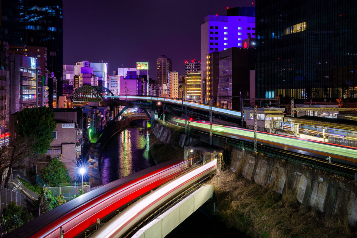 Elevated train lines in Tokyo at night, with light trails reflecting on a canal below and illuminated buildings in the background.