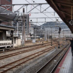 Multiple sets of railway tracks converge toward the distance beneath a grid of steel catenary supports. A man in a dark suit walks along the yellow tactile line on Platform 4, while another commuter waits across the tracks. Overhead signs mark platform numbers, and signal lights display red and amber. The station canopy and surrounding buildings form an industrial backdrop, with a faint view of hills beyond the city.