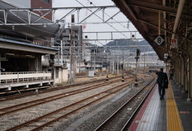 Multiple sets of railway tracks converge toward the distance beneath a grid of steel catenary supports. A man in a dark suit walks along the yellow tactile line on Platform 4, while another commuter waits across the tracks. Overhead signs mark platform numbers, and signal lights display red and amber. The station canopy and surrounding buildings form an industrial backdrop, with a faint view of hills beyond the city.