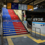 A color-coded stairway inside Nippori Station directs passengers to the Keisei Line platforms, the primary transfer point for travelers heading to Narita Airport on the high-speed Skyliner. Nippori serves as a major interchange between JR East lines, the Keisei Main Line, and the Nippori–Toneri Liner, linking northern Tokyo neighborhoods to the broader regional network. The mixed architecture—older steel passageways alongside newer safety upgrades like platform-edge barriers—reflects the station’s layered development since its opening in 1905. Today, Nippori remains one of Tokyo’s most important cross-rail transfer nodes, especially for airport-bound passengers navigating the city’s extensive transit system.