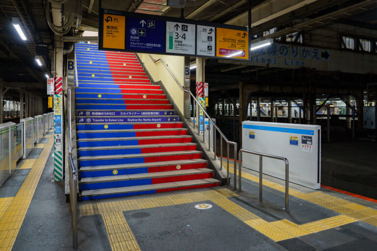 Transfer Stairway to the Keisei Line at Nippori Station 1 A color-coded stairway inside Nippori Station directs passengers to the Keisei Line platforms, the primary transfer point for travelers heading to Narita Airport on the high-speed Skyliner. Nippori serves as a major interchange between JR East lines, the Keisei Main Line, and the Nippori–Toneri Liner, linking northern Tokyo neighborhoods to the broader regional network. The mixed architecture—older steel passageways alongside newer safety upgrades like platform-edge barriers—reflects the station’s layered development since its opening in 1905. Today, Nippori remains one of Tokyo’s most important cross-rail transfer nodes, especially for airport-bound passengers navigating the city’s extensive transit system.