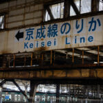 A weathered overhead passage at Nippori Station directs passengers toward the Keisei Line, one of Tokyo’s key links to Narita Airport via the Skyliner service. Nippori has served as a major interchange since the early 20th century, connecting JR East’s Yamanote, Keihin-Tōhoku, and Jōban lines with the private Keisei network.

The aging steelwork and faded signage reflect the layered construction of the station complex, where older elevated structures remain in daily use alongside modernized ticketing halls and platform edge barriers. Despite its utilitarian appearance, this transfer point moves thousands of airport-bound travelers each day, making it one of Tokyo’s busiest cross-rail connections.