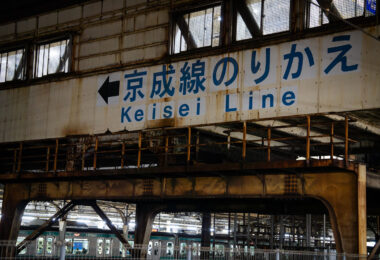 A weathered overhead passage at Nippori Station directs passengers toward the Keisei Line, one of Tokyo’s key links to Narita Airport via the Skyliner service. Nippori has served as a major interchange since the early 20th century, connecting JR East’s Yamanote, Keihin-Tōhoku, and Jōban lines with the private Keisei network.

The aging steelwork and faded signage reflect the layered construction of the station complex, where older elevated structures remain in daily use alongside modernized ticketing halls and platform edge barriers. Despite its utilitarian appearance, this transfer point moves thousands of airport-bound travelers each day, making it one of Tokyo’s busiest cross-rail connections.