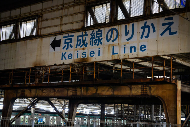 Keisei Line Transfer Passage at Nippori Station 1 A weathered overhead passage at Nippori Station directs passengers toward the Keisei Line, one of Tokyo’s key links to Narita Airport via the Skyliner service. Nippori has served as a major interchange since the early 20th century, connecting JR East’s Yamanote, Keihin-Tōhoku, and Jōban lines with the private Keisei network.
The aging steelwork and faded signage reflect the layered construction of the station complex, where older elevated structures remain in daily use alongside modernized ticketing halls and platform edge barriers. Despite its utilitarian appearance, this transfer point moves thousands of airport-bound travelers each day, making it one of Tokyo’s busiest cross-rail connections.