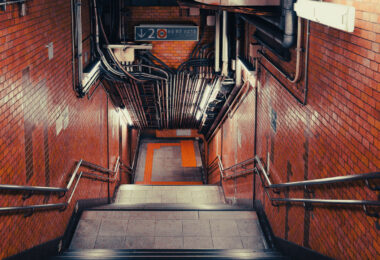 A passageway leading down to the Tokyo Metro Marunouchi Line, part of one of Tokyo’s oldest surviving subway corridors. The Marunouchi Line began opening in stages between 1954 and 1962, making it the second-oldest subway line in the city after the Ginza Line. Many stations along the route—especially the early central segments—retain architectural and material choices from Japan’s postwar reconstruction period, including ceramic wall tile, exposed conduit, and compact stairways built before modern accessibility standards were introduced.

The dense overhead wiring found in older Marunouchi Line interchanges reflects decades of retrofits to accommodate improved lighting, ventilation, fire-suppression systems, and communication networks, all integrated into the original structural envelope. These spaces remain essential transfer points connecting central Tokyo’s commercial districts with major JR East hubs such as Shinjuku, Tokyo Station, and Ochanomizu.

Although the Marunouchi Line has undergone rolling upgrades—including new rolling stock (02 series replacements), platform screen doors, and improved wayfinding—the underlying layout in many stations still conveys the functional, space-efficient design philosophy of mid-20th-century Japanese subway engineering.