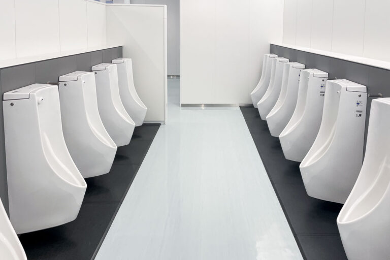 Symmetry in Clean Design 1 A perfectly symmetrical row of urinals inside a modern public restroom in Japan, where minimalist design meets meticulous cleanliness. The glossy white fixtures and pale reflective flooring emphasize balance and precision, creating an unexpectedly architectural sense of order in a utilitarian space. The visual rhythm of repetition — identical forms mirrored across the corridor — captures Japan’s cultural devotion to hygiene, geometry, and quiet functionality in even the most mundane public settings.