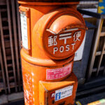 A cylindrical orange post box on a street in Kyoto, photographed in March 2023. This style of mailbox—introduced in the early 1900s during Japan’s push to modernize its postal infrastructure—became a familiar feature across the country through the mid-20th century. Its rounded form and front mail slot were designed to be durable, weather-resistant, and easy to recognize even in densely built neighborhoods.

Although newer rectangular mailboxes are now common in urban areas, many historic districts in Kyoto retain these older designs to preserve the traditional streetscape. Today, they continue to function as part of Japan Post’s network, blending everyday utility with a tangible link to the city’s architectural and civic history.