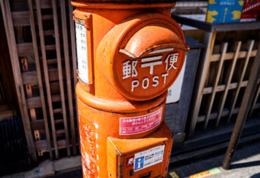 A cylindrical orange post box on a street in Kyoto, photographed in March 2023. This style of mailbox—introduced in the early 1900s during Japan’s push to modernize its postal infrastructure—became a familiar feature across the country through the mid-20th century. Its rounded form and front mail slot were designed to be durable, weather-resistant, and easy to recognize even in densely built neighborhoods.

Although newer rectangular mailboxes are now common in urban areas, many historic districts in Kyoto retain these older designs to preserve the traditional streetscape. Today, they continue to function as part of Japan Post’s network, blending everyday utility with a tangible link to the city’s architectural and civic history.